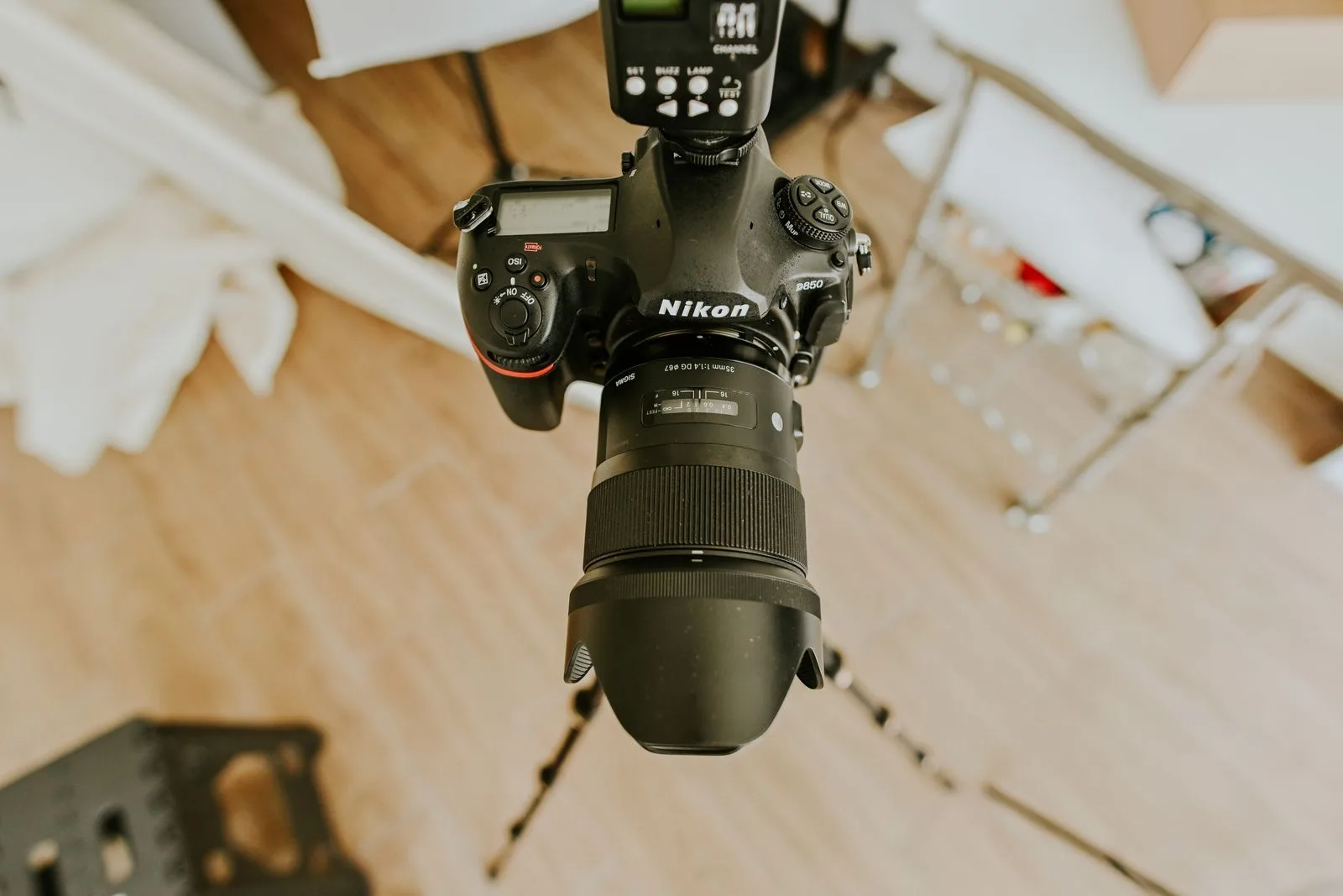A DSLR on a tripod with a fast prime lens and hood, standing on pale wood flooring in a studio with light stands blurred behind it.