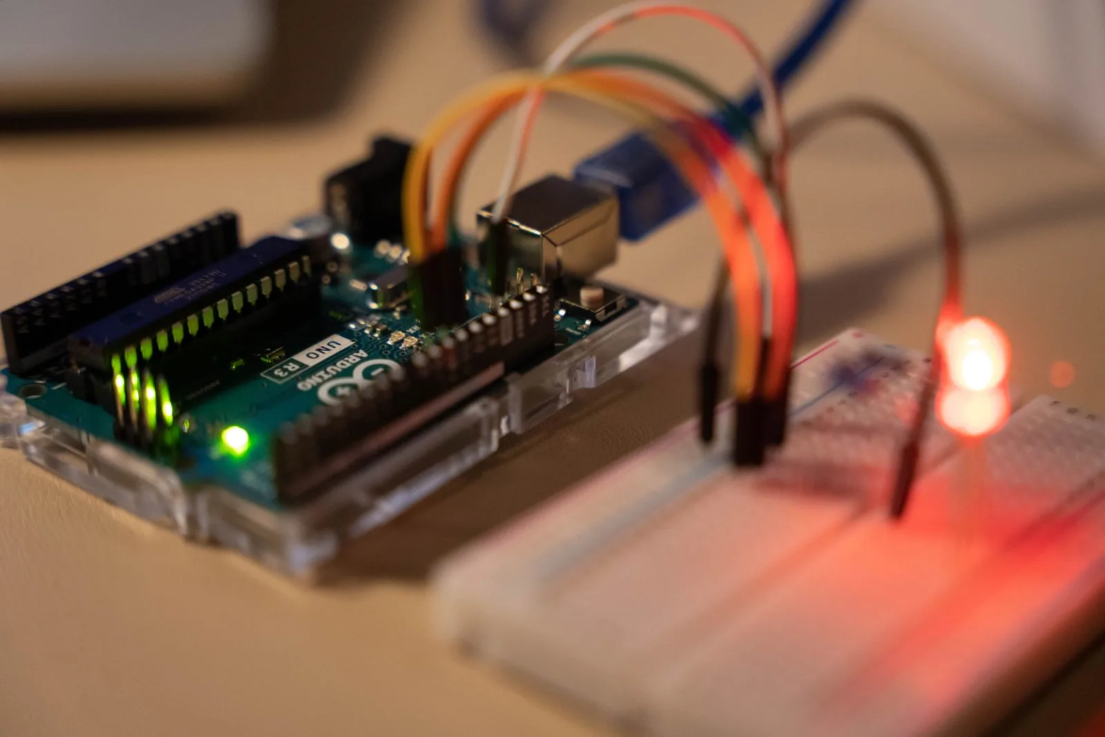 Close-up of an Arduino board on a desk with orange, white and green jumper wires arcing from its header pins to a white breadboard where a single red LED is lit.