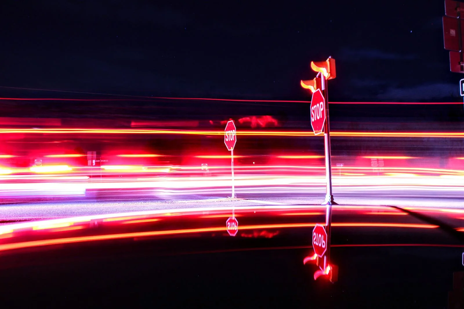 A night-time long exposure at a road intersection: two red-and-white stop signs stand lit against a black sky while red and white light trails streak horizontally across the frame behind them.