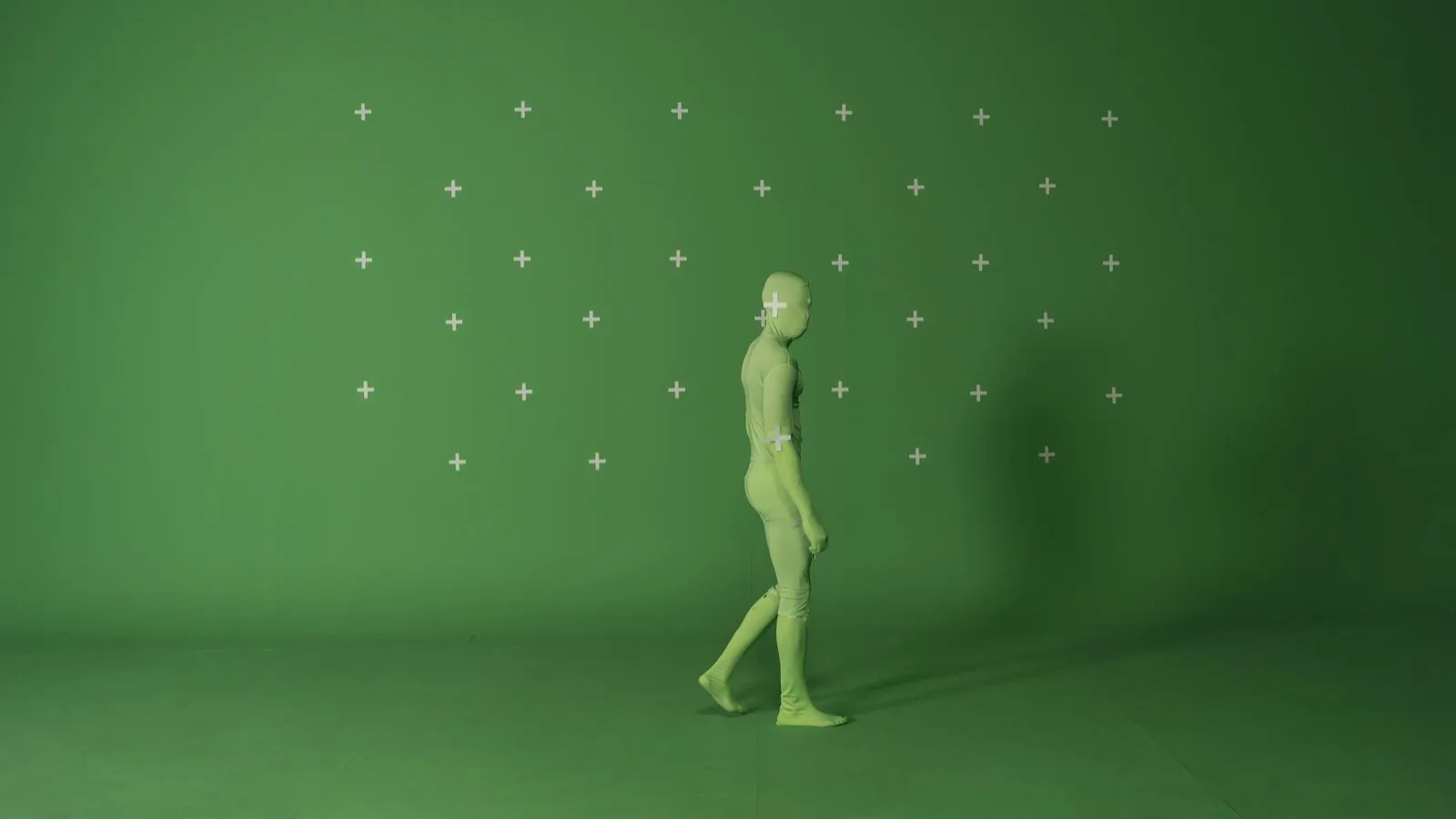 A figure in a motion-capture suit marked with tracking dots, walking against a green screen.