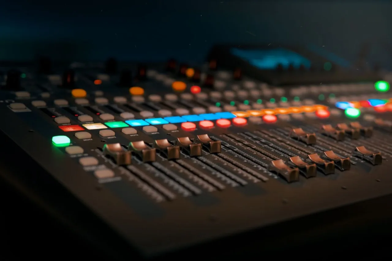 A darkened mixing console, its channel buttons lit in amber and cyan, faders running out of focus.