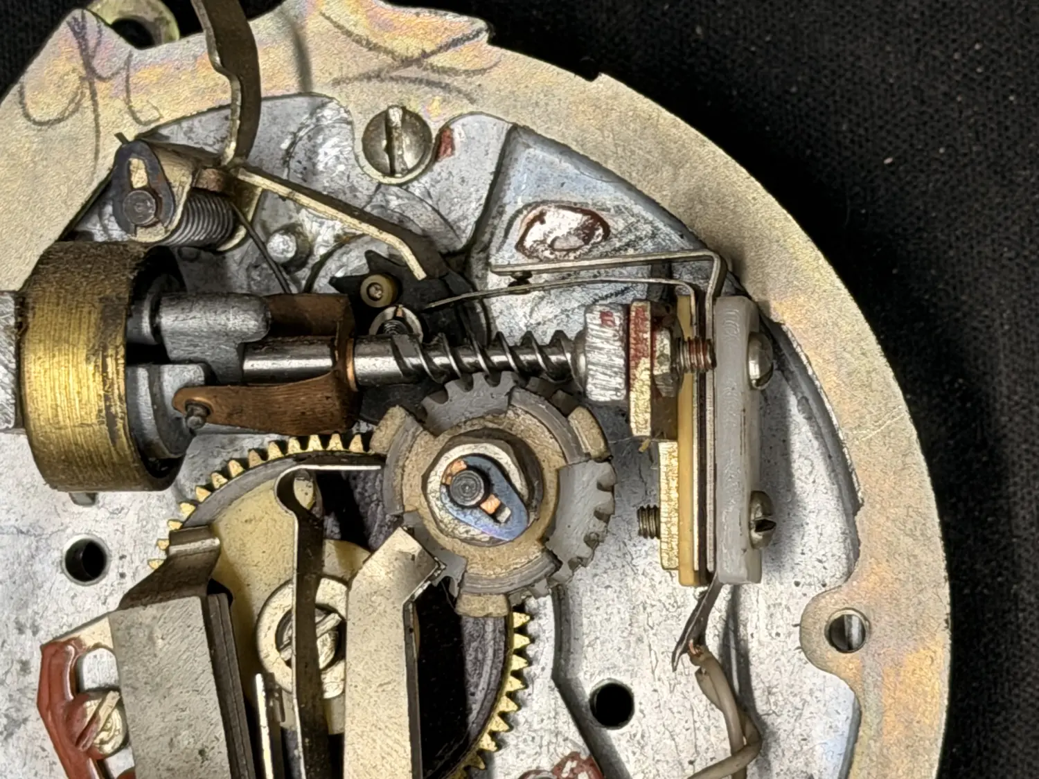 Macro view of the rotary dial mechanism removed from the phone: a brass gear train, a governor, coiled springs and a set of contact points mounted on a circular alloy plate, all lightly tarnished.