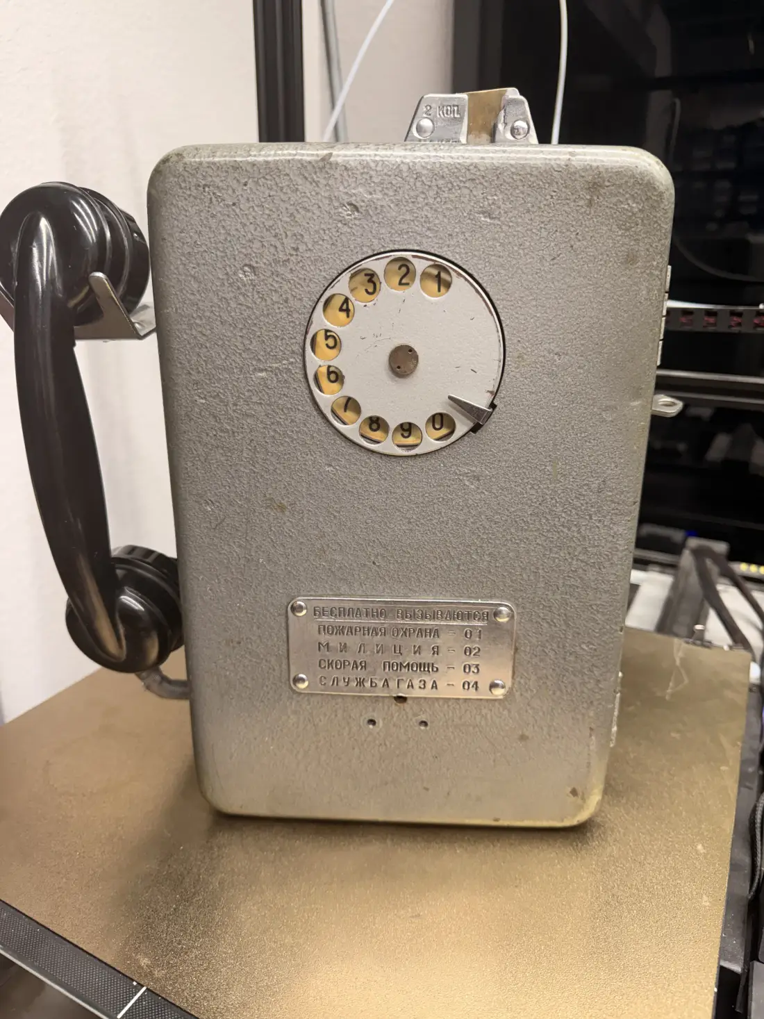 The front of the AMT-69: pebbled grey enamel, a rotary dial with Cyrillic-era numbering, a black handset hanging on the left, and a metal plate listing the free emergency numbers — fire 01, police 02, ambulance 03, gas service 04.