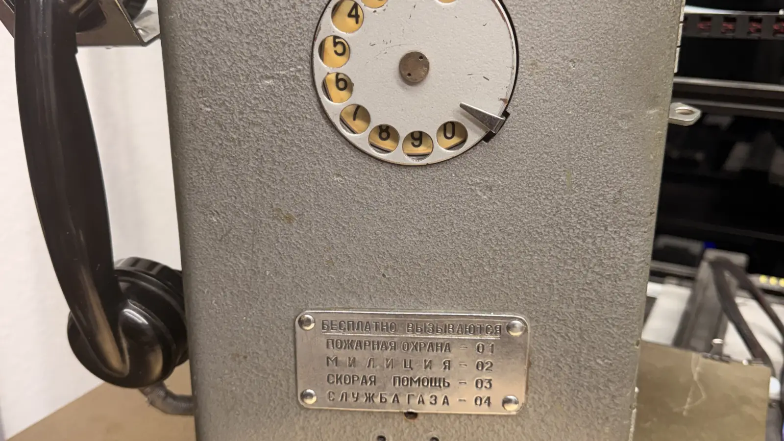 The face of a Soviet AMT-69 payphone: a rotary dial above a metal plate listing emergency numbers in Cyrillic, with a black handset hanging beside it.