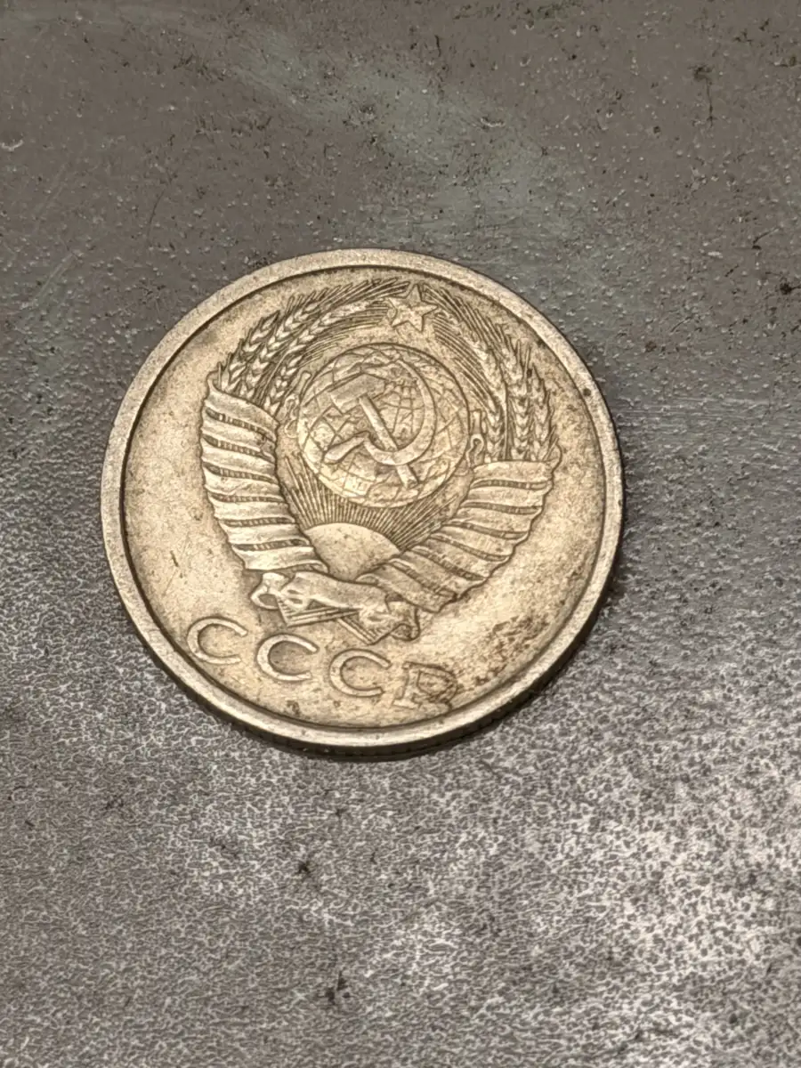 A worn Soviet kopeck coin resting on the grey enamel of the phone, its face showing the USSR state emblem — a hammer and sickle over a globe, framed by wheat ears — and the letters СССР.
