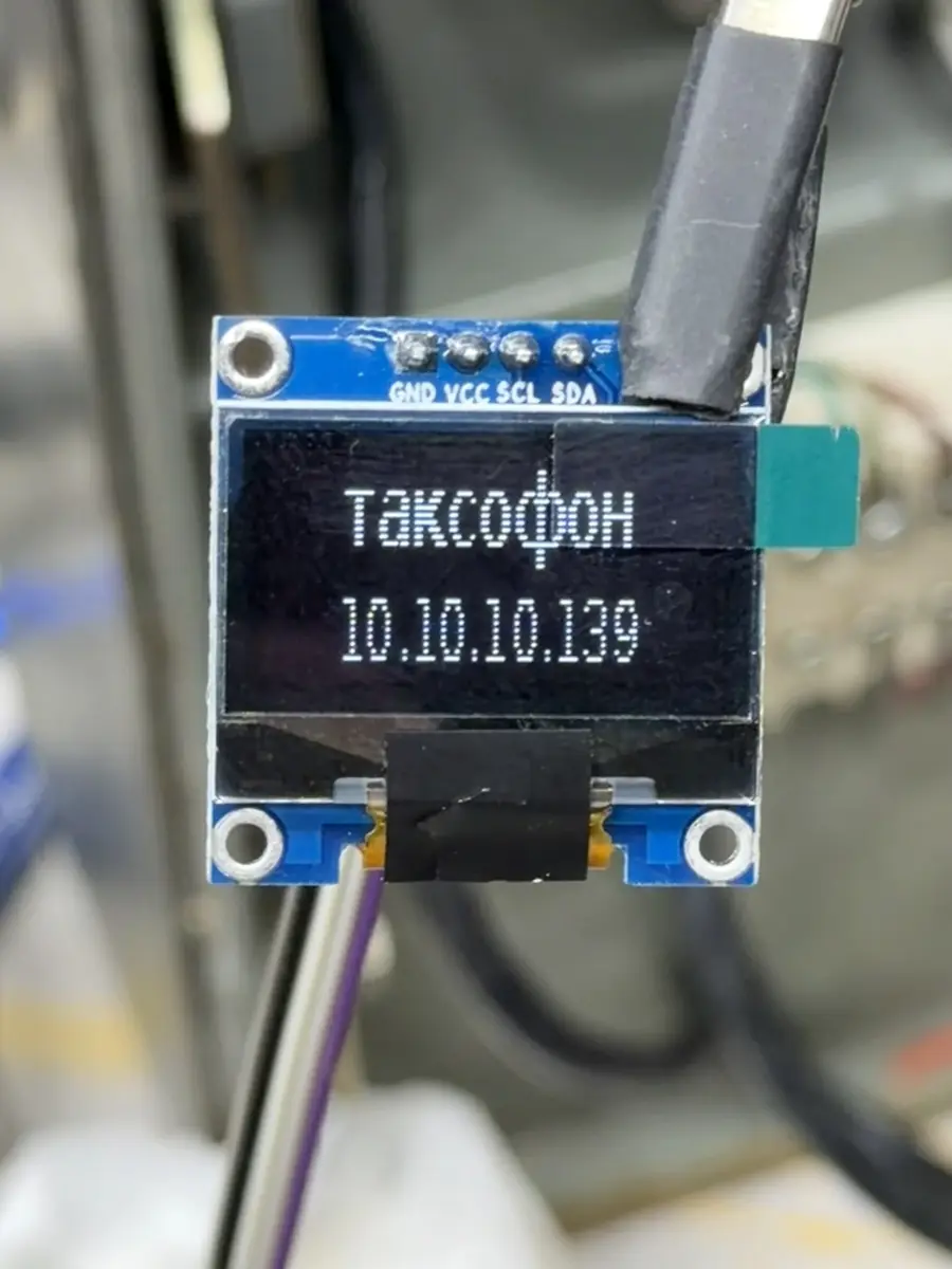 A small blue I2C OLED module held up in front of the open phone, its screen reading Таксофон above the address 10.10.10.139.