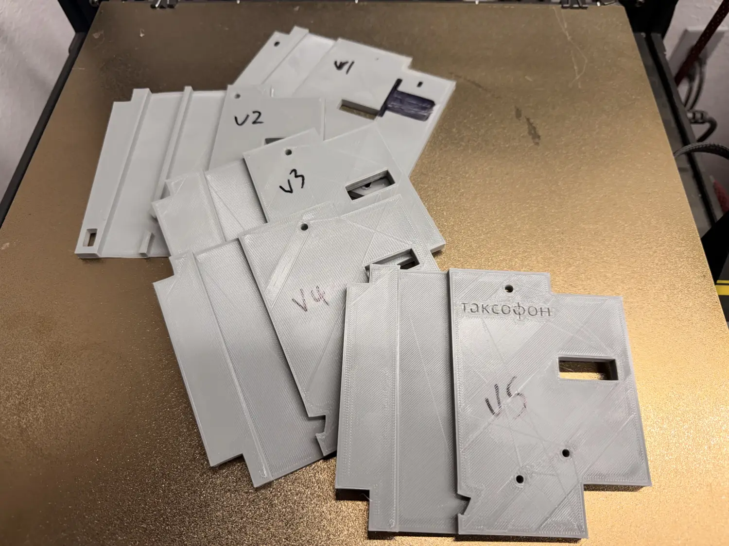 Six 3D-printed versions of the coin mechanism fanned out on a printer bed, five of them hand-labelled in marker V1 through V5, and the sixth carrying the embossed word Таксофон instead of a number.